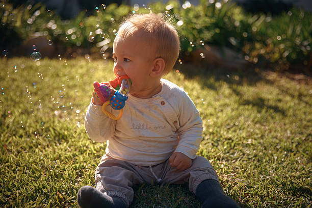 A sweet photo of a baby sitting in the garden at home, chewing on a toy while bubbles float around him. A playful and gentle moment filled with curiosity and springtime charm.