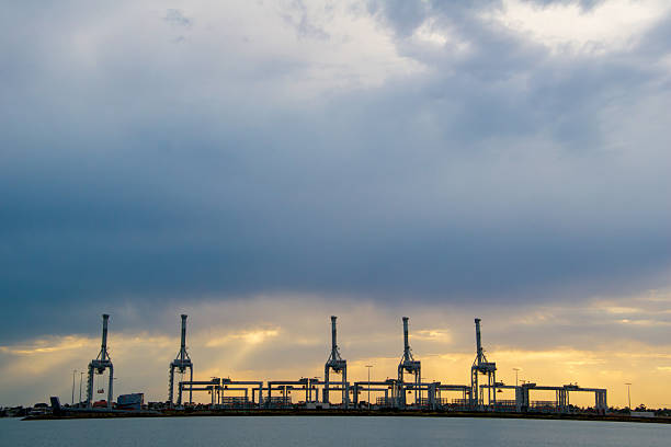 Industrial port cranes silhouetted against a dramatic, cloud-filled sky at sunset