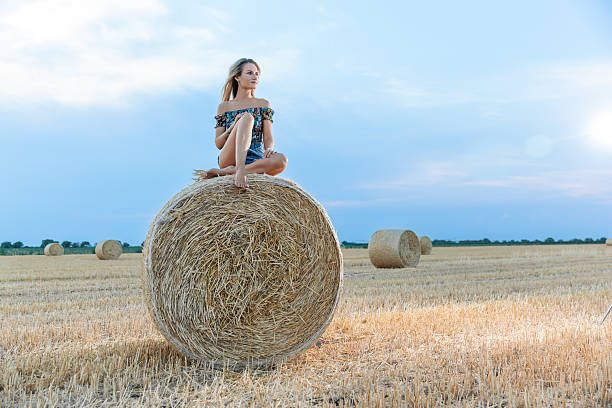 istock Young woman sitting on a round haystack in a mowed field 2231309011