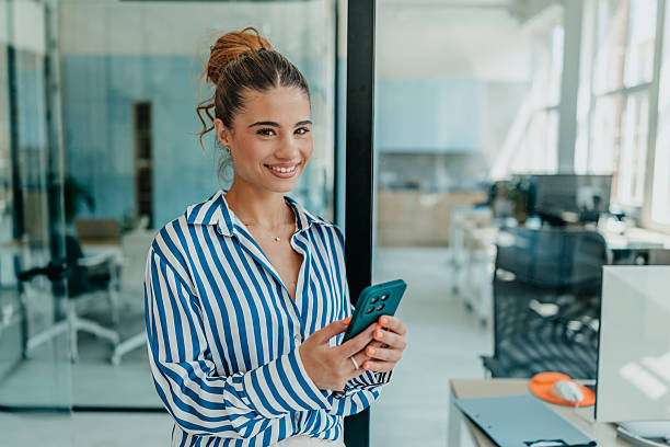 istock Cheerful young woman smiling while using smartphone in bright office during daytime 2231310141