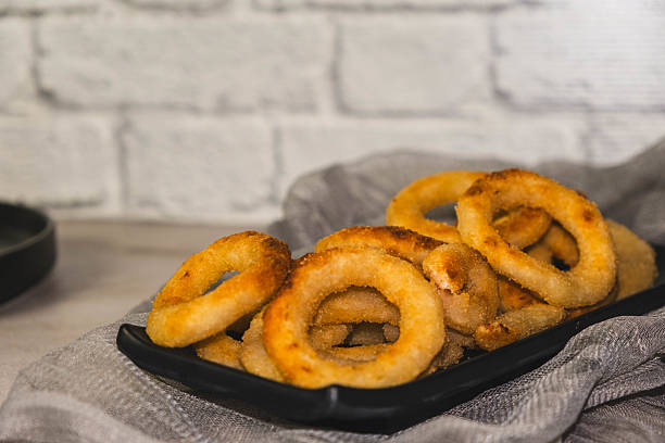 A plate of fried onion rings sits on a table. The rings are golden brown and appear to be crispy. The plate is black and the table is white