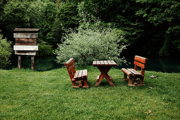 Wooden benches and a table stand on green grass by a calm river, surrounded by dense trees. A rustic signpost adds charm to the secluded spot. Krupajsko vrelo in Serbia