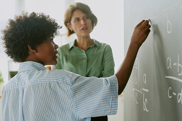 istock Teenage Black Boy Solving Math Problem on Chalkboard with Teacher Observing 2231335069