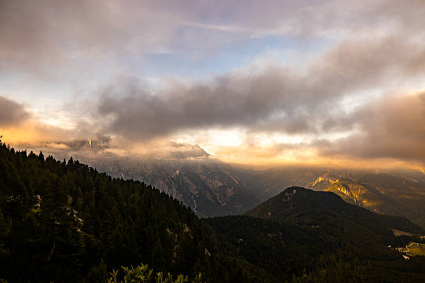 Scenic mountain valley view with low clouds illuminated by golden sunlight. Forested hills and rugged alpine peaks appear through the mist during sunrise or sunset.