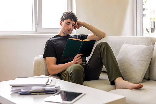 istock Young male reading book on sofa 2231347337