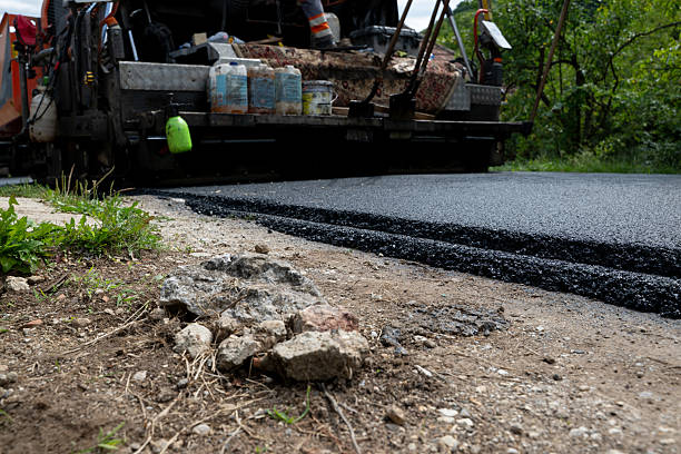 A paving machine spreads a fresh layer of asphalt on a rural road, with construction tools and containers visible on the equipment. The low-angle perspective highlights the contrast between rough ground and the smooth, newly laid blacktop, emphasizing infrastructure development and road improvement.
