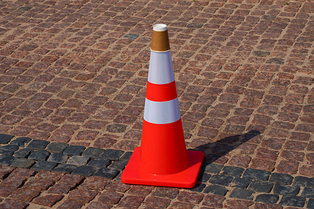 istock Bright orange traffic cone with white reflective stripes placed on a cobblestone pavement. A small cardboard cup is placed upside down on the top of the cone, creating an unusual and humorous urban scene. 2231361664