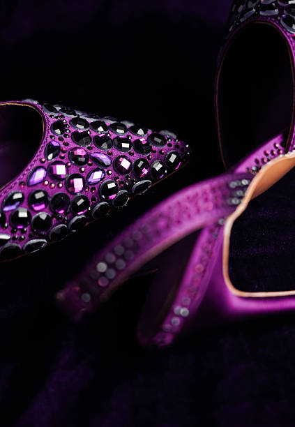 Close-up view of a pair of elegant purple high-heeled shoes adorned with numerous sparkling crystals and gemstones. The shoes rest on a rich, dark purple velvet background, creating a dramatic and luxurious contrast.  The focus is primarily on the intricate jeweled detailing on the shoes, with a shallow depth of field blurring the background and parts of the shoe. The lighting accentuates the shimmer and shine of the crystals.