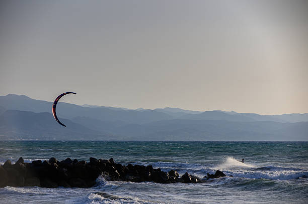 Scenic view of a kitesurfer riding waves near rocky shore with mountains in the background.