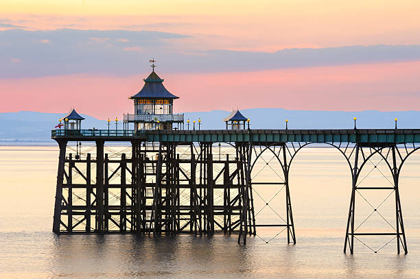 A serene view of Clevedon Pier in Somerset, England, at dusk. The pier's Victorian architecture, including its ornate pavilion, stands silhouetted against a pastel sky, offering a glimpse into the town's seaside heritage and the enduring appeal of coastal landmarks.