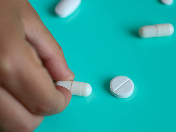 ​A human hand gently takes a white capsule from a turquoise background, surrounded by other white pills. The shot conveys the idea of choice, health care, and personal treatment, perfectly suited for illustrating medical themes.