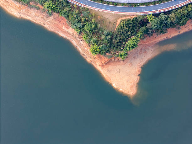 This aerial photograph captures a winding road curving gracefully along the edge of a serene lake, surrounded by lush greenery and terraced fields. The visual style is naturalistic, emphasizing the harmony between human-made structures and the environment. The image conveys tranquility and could be used for travel, nature, or infrastructure-related purposes.