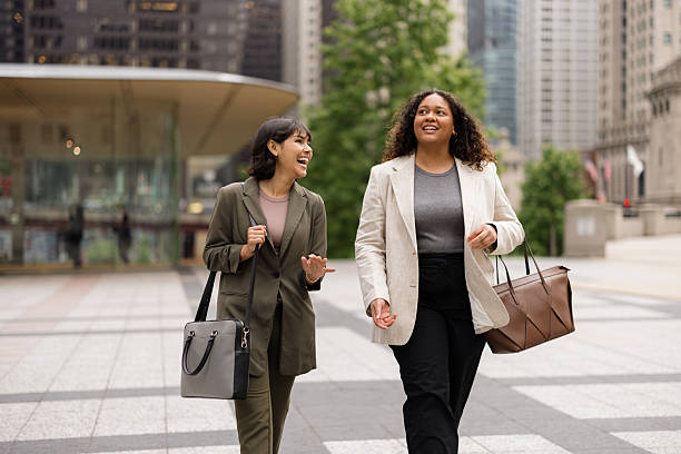 Two young businesswomen walking and chatting on a bustling Chicago street, enjoying the vibrant city atmosphere and each other's company