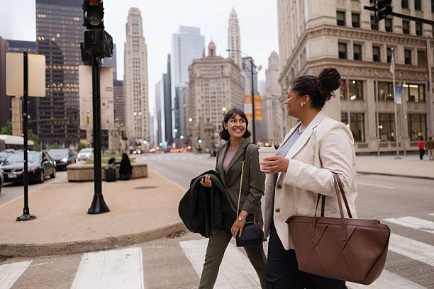 Two businesswomen are walking and talking on a crosswalk in Chicago, holding coffee cups and bags