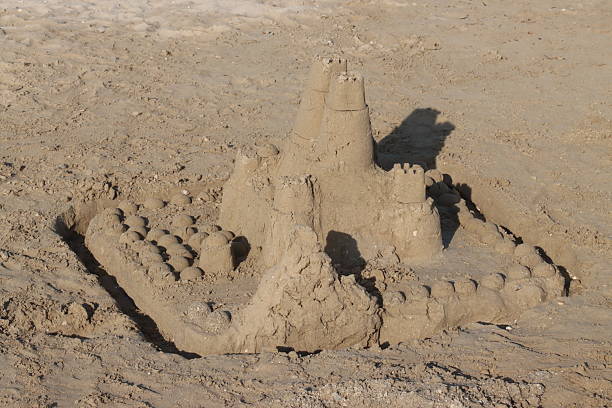 Stock photo showing a man building a complex sandcastle built on the beach in Goa, India. This structure was made with a bucket and green spade, with an exterior wall modification to turn it into a fortress