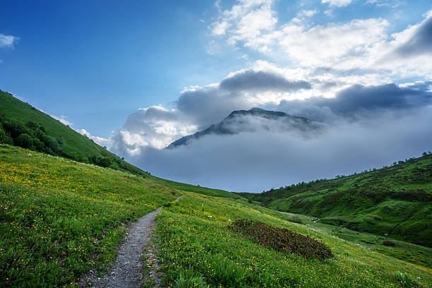 A breathtaking view of a mountain valley featuring mist-shrouded peaks, lush meadows, and a winding footpath leading through the tranquil greenery under a bright blue sky.