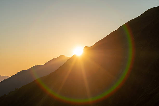 Sunrise peeking over a mountain range, casting light and shadows with a colorful lens flare.