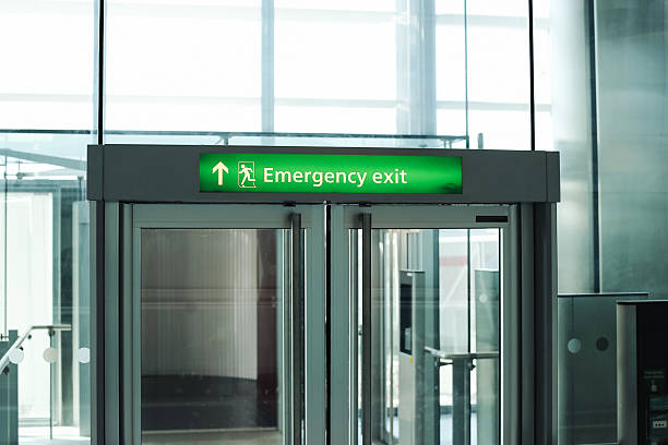 istock Emergency exit door in modern public building interior. Green safety sign with arrow and running man symbol above transparent glass door. 2231410569