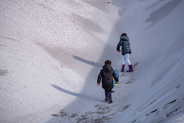 Two children are walking down a sandy slope on a winter beach, carrying toys and wearing warm winter clothes