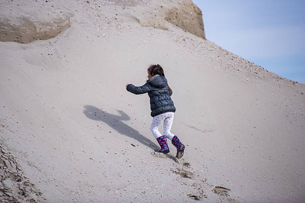 Young girl running down a steep sand dune on a sunny beach, wearing a warm winter jacket and boots