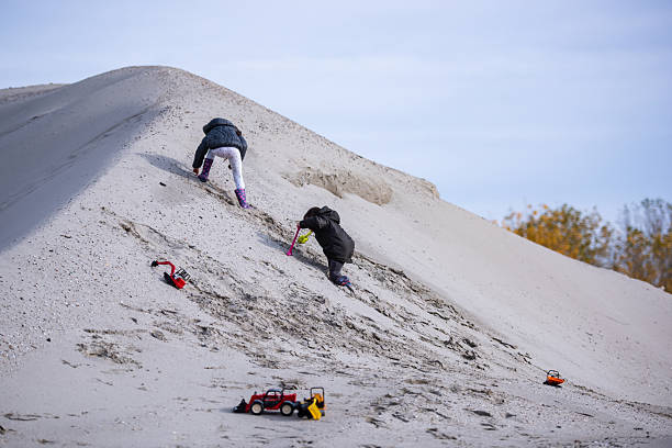 Two children climbing a sand dune, joyfully playing with toy trucks under a bright autumn sun, embracing the spirit of adventure and fun