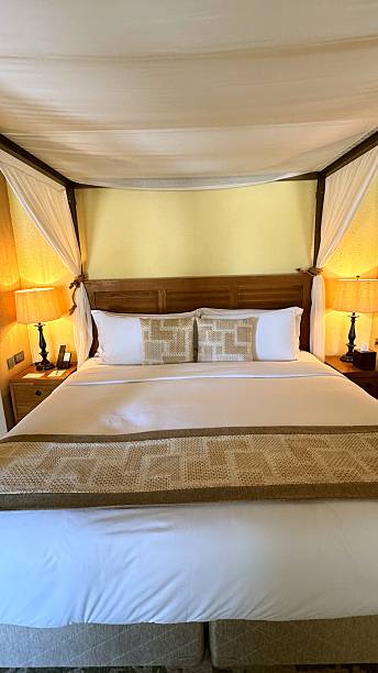 istock Canopy bed with white sheets, pillows and decorative bed runner illuminated by two lamps on wooden nightstands in hotel room with yellow wall 2231413353
