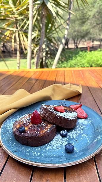 Wooden table setting featuring golden French toast topped with fresh berries, dusted with powdered sugar, blurred garden landscape behind