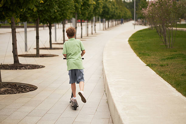 istock Caucasian Boy Riding Scooter Along Urban Park Pathway on Summer Day 2231417765