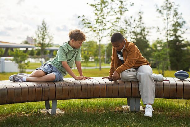 Group of multiracial children smiling, sitting at stone chair, playing together outdoors in summer park. Adorable kids have fun playing in playground. Selective focus on Asian girl