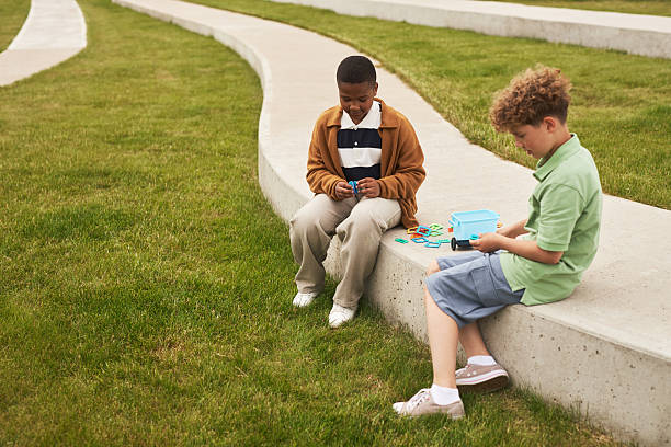 Black boy and Caucasian boy sitting on curved concrete bench playing with colorful plastic toys, both children focused on activity, outdoor setting with copy space