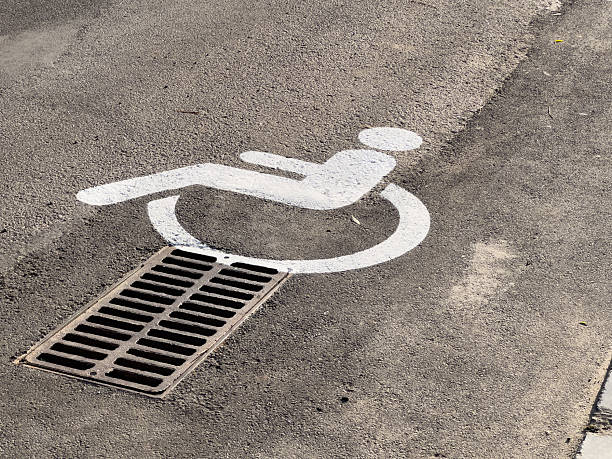 A white accessibility symbol is painted on pavement, positioned directly over a metal drainage grate in an urban setting