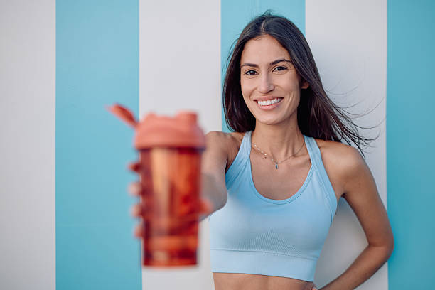 Smiling sportswoman proudly holding a protein shake against a vibrant blue and white background, radiating health and vitality