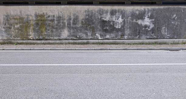 istock Dirty plaster wall almost fully covered with moss and stains. Sidewalk and asphalt road in front 2231442549