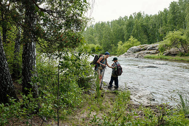 Woman assisting boy crossing shallow river in forest, both carrying backpacks and hiking gear, surrounded by lush green trees and rocky terrain