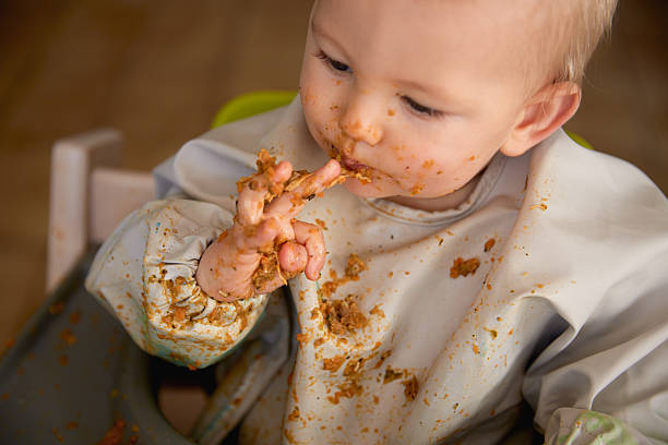 A cute and candid photo of a baby self-feeding with his fingers, food smeared everywhere and a full bib catching the mess. A joyful and chaotic moment capturing the fun of learning to eat.