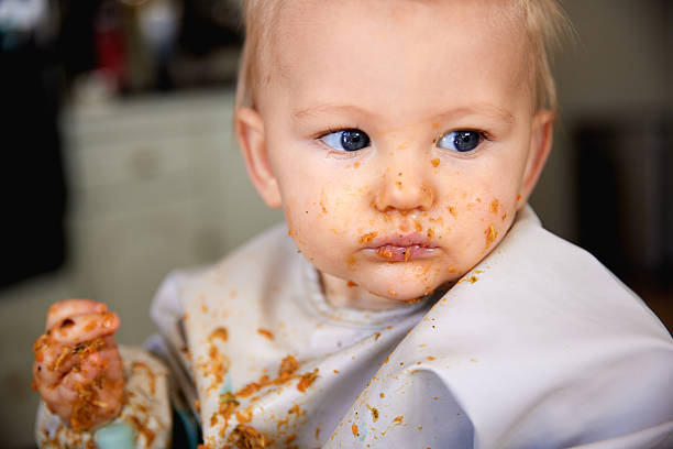 A cute and candid photo of a baby self-feeding with his fingers, food smeared everywhere and a full bib catching the mess. A joyful and chaotic moment capturing the fun of learning to eat.