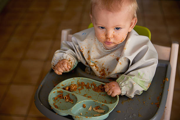 A cute and candid photo of a baby self-feeding with his fingers, food smeared everywhere and a full bib catching the mess. A joyful and chaotic moment capturing the fun of learning to eat.