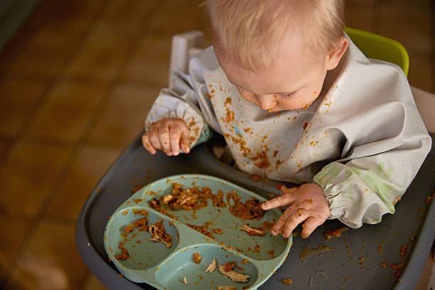 A cute and candid photo of a baby self-feeding with his fingers, food smeared everywhere and a full bib catching the mess. A joyful and chaotic moment capturing the fun of learning to eat.