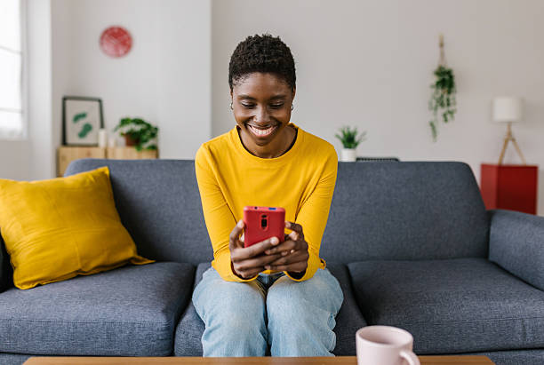 istock Happy young african woman using mobile phone sitting on sofa at home 2231456280