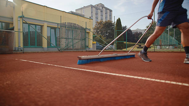 A tennis player is scrubbing a clay court after practice using a large brush. The player is wearing athletic clothing and is maintaining the clay surface in an outdoor sports facility featuring surrounding greenery and urban buildings.