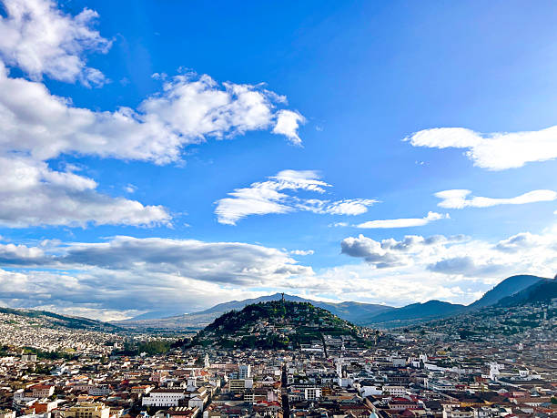 Quito's cityscape stretches beneath a dynamic sky, with the iconic El Panecillo hill standing prominently against a backdrop of rolling mountains
