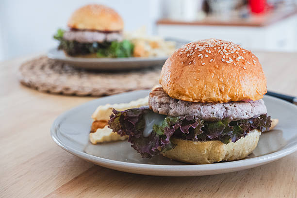 Close-up of a delicious plant-based burger with red leaf lettuce and crinkle-cut fries on a white plate set on a wooden table, with another blurred burger in the background.