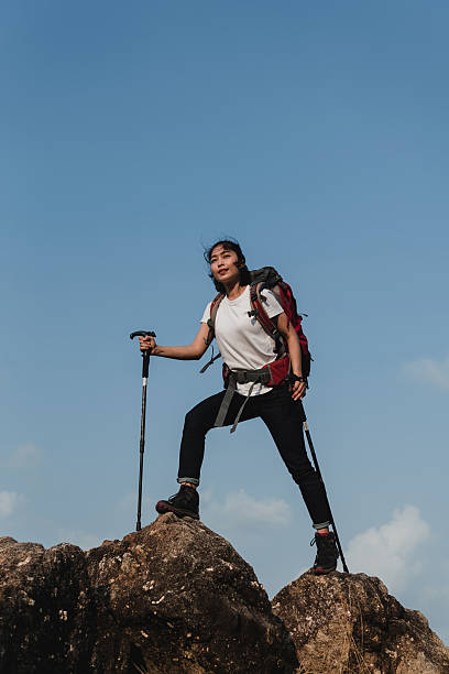 Confident Asian female hiker with a red backpack and trekking poles standing triumphantly on a rocky mountain peak under a clear blue sky.