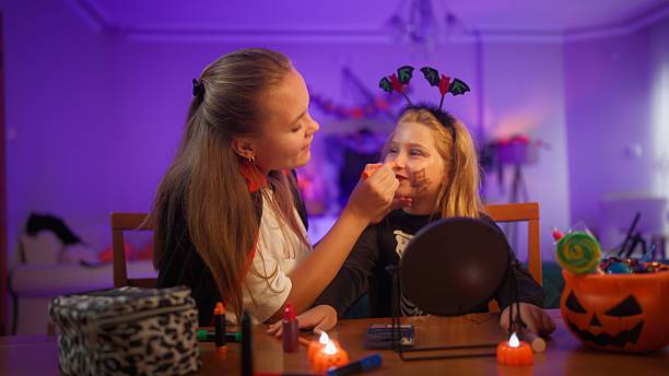 A mother is putting on Halloween make up on her daughter's face in the living room at home.