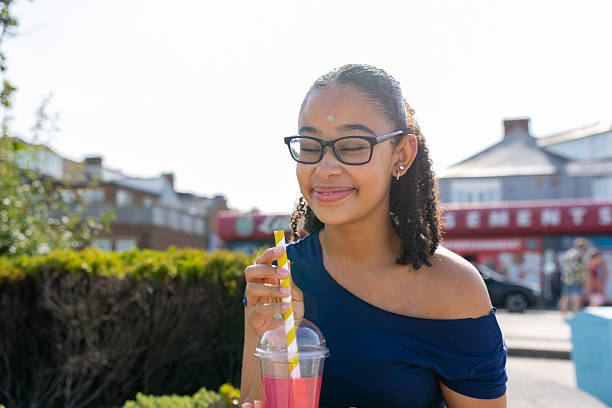 A close-up shot of a happy teenage girl sitting outside under the sunlight, smiling with her eyes closed while holding a refreshing fruit beverage with a striped straw. Natural candid summer lifestyle portrait.Videos are available similar to this scenario.