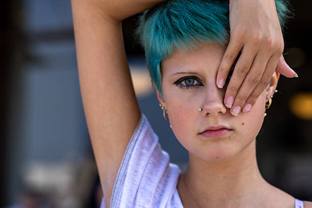 A close-up portrait shot of a teenage girl with short green-dyed hair standing outdoors, looking directly at the camera while holding her hand over one of her eyes. She is wearing a casual white T-shirt.Videos are available similar to this scenario.