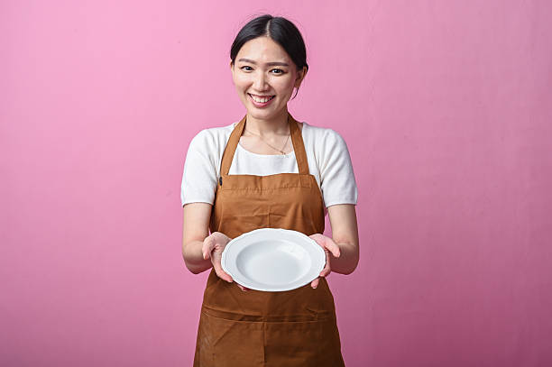istock Asian woman in a brown apron holding a clean white ceramic plate with both hands, smiling warmly against a plain pink background 2231491624