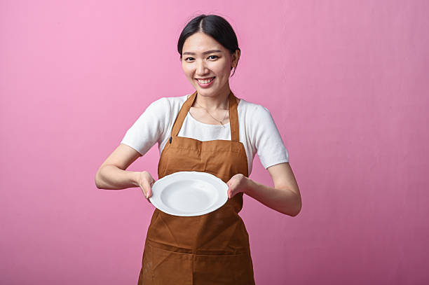 istock Asian woman in a brown apron holding a clean white ceramic plate with both hands, smiling warmly against a plain pink background 2231491679