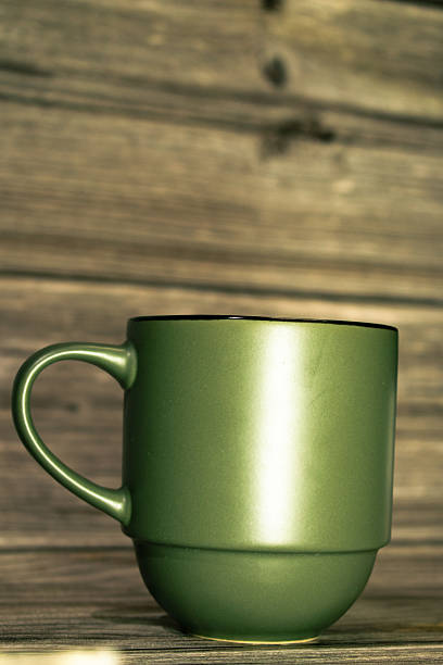 A green ceramic mug filled with a dark beverage, possibly coffee, sits on a rustic wooden surface. The mug has a simple, classic shape with a smooth finish. The wood grain beneath it suggests a natural, cozy setting, enhancing a warm and inviting atmosphere.