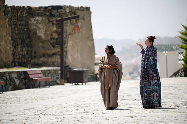 Full length view of two women in abayas exploring the sunny courtyard of Shamsan Ottoman Castle, walking and talking with historic stone walls in the distance.